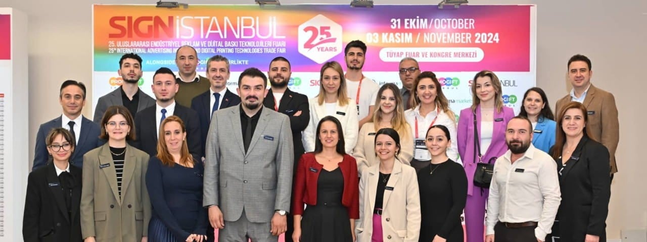 Ekibin, etkinlik tabelası önünde iş kıyafetleriyle  birlikte çekilmiş grup fotoğrafı.
